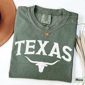 Puede incluir: Una camiseta verde con la palabra "TEXAS" en letras blancas y una silueta blanca de un toro longhorn. La camiseta tiene un aspecto desgastado y est&aacute; hecha de una tela suave y c&oacute;moda.