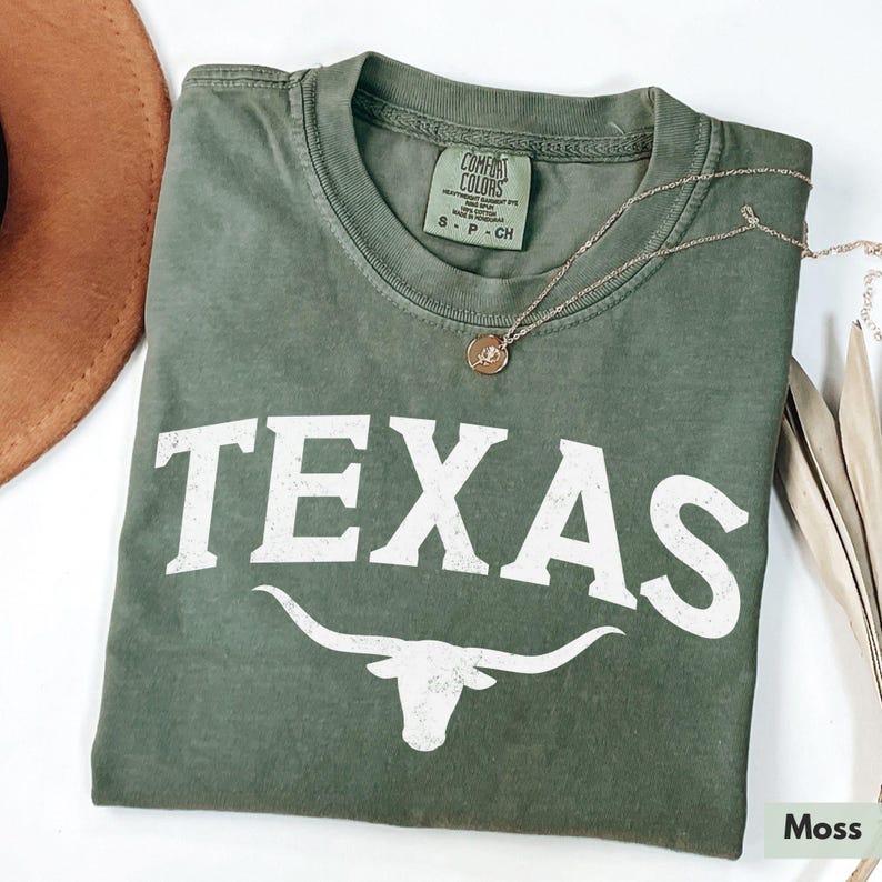 Puede incluir: Una camiseta verde con la palabra "TEXAS" en letras blancas y una silueta blanca de un toro longhorn. La camiseta tiene un aspecto desgastado y est&aacute; hecha de una tela suave y c&oacute;moda.