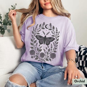May include: A lavender-colored t-shirt with a graphic of a black butterfly in a floral wreath with a crescent moon and stars. The shirt is made of a soft, comfortable material.