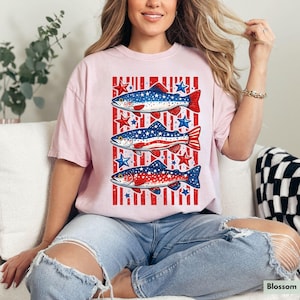 May include: A light pink t-shirt featuring a patriotic design with three fish. Each fish is decorated with red, white, and blue colors, and surrounded by stars. The shirt has a casual, relaxed fit.