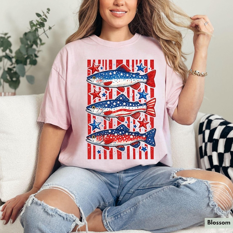 May include: A light pink t-shirt featuring a patriotic design with three fish. Each fish is decorated with red, white, and blue colors, and surrounded by stars. The shirt has a casual, relaxed fit.