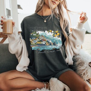 以下が含まれることがあります： イタリアのアマルフィ海岸のグラフィックプリントが施されたダークグレーのTシャツ。グラフィックは、建物、ボート、レモンのカラフルなイラストが特徴です。グラフィックの下には「Amalfi Coast, Italy」というテキストが印刷されています。