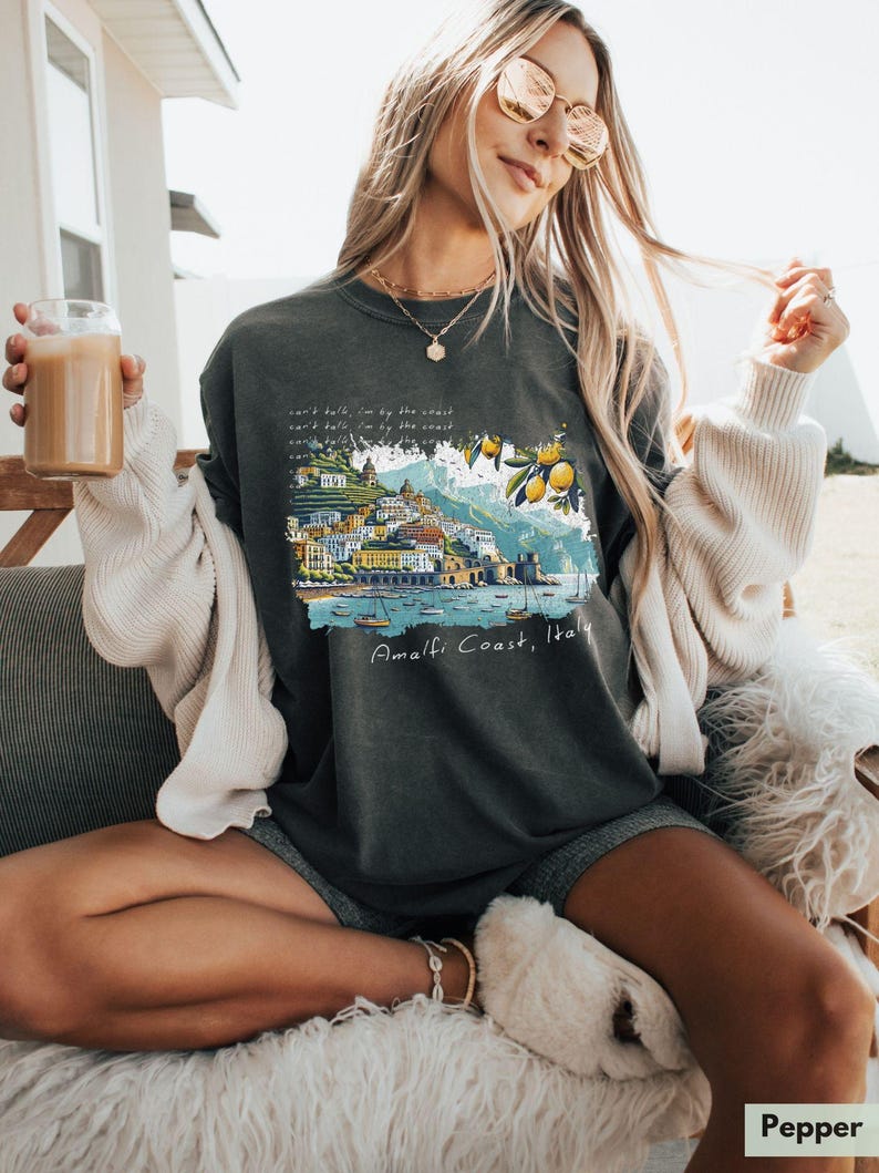 以下が含まれることがあります： イタリアのアマルフィ海岸のグラフィックプリントが施されたダークグレーのTシャツ。グラフィックは、建物、ボート、レモンのカラフルなイラストが特徴です。グラフィックの下には「Amalfi Coast, Italy」というテキストが印刷されています。