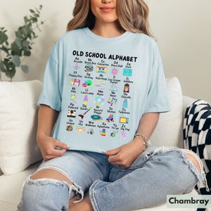 May include: A light blue t-shirt with a graphic of an alphabet printed on it. The graphic features a different 80s themed icon for each letter of the alphabet. The text "OLD SCHOOL ALPHABET" is printed at the top of the graphic.