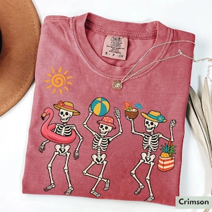 May include: A crimson red t-shirt with a graphic of three skeletons wearing summer accessories. The skeletons are holding a beach ball, a pineapple, and a tropical drink. The text "COMFORT COLORS" is visible on the tag.