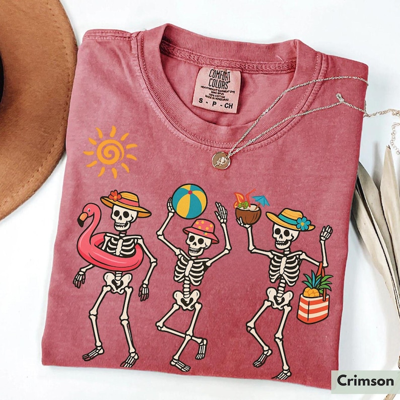 May include: A crimson red t-shirt with a graphic of three skeletons wearing summer accessories. The skeletons are holding a beach ball, a pineapple, and a tropical drink. The text "COMFORT COLORS" is visible on the tag.