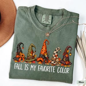 May include: A moss green t-shirt featuring a graphic of five distinct witch hats decorated with autumn leaves, sunflowers, and berries. The text "FALL IS MY FAVORITE COLOR" is printed in white below the hats. A thin gold necklace rests on the collar.
