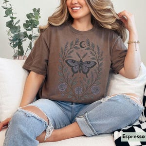 May include: A brown t-shirt with a floral design featuring a butterfly and a crescent moon. The shirt is worn by a person with long brown hair and is paired with blue jeans.