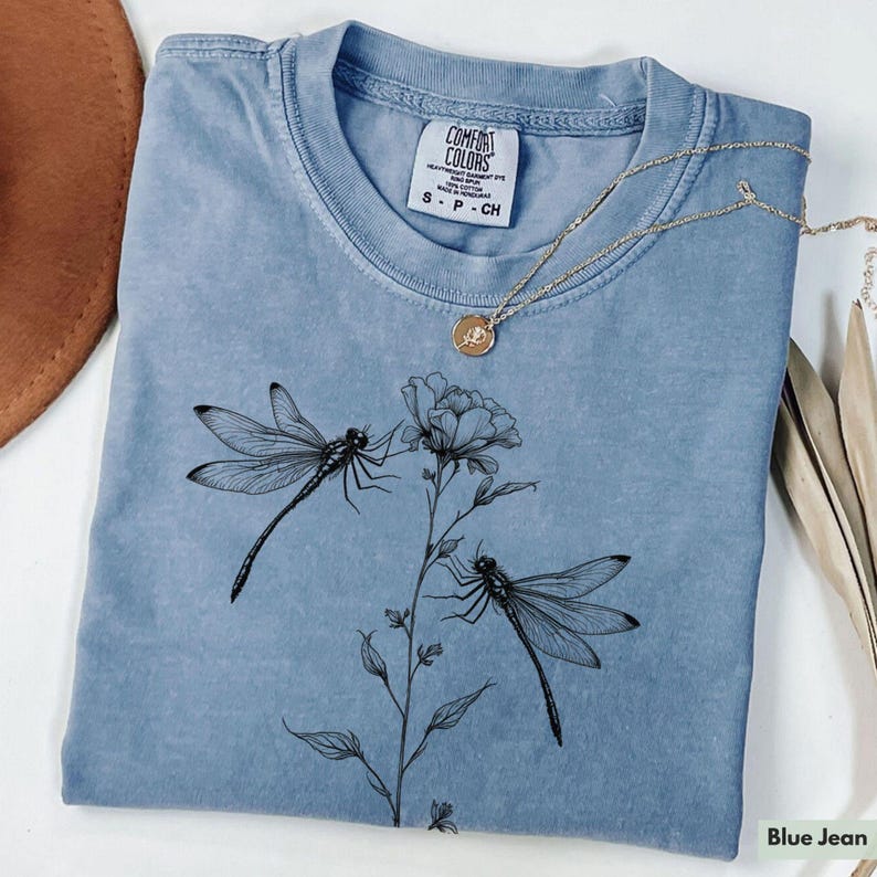 May include: A blue jean t-shirt with a floral design featuring two dragonflies and the text "Comfort Colors" in white.