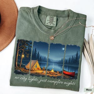 Op de afbeelding: Een groen T-shirt met een afbeelding van een camping in de nacht. De afbeelding bevat een tent, een kampvuur, een kano en bomen. De tekst "no city lights, just campfire nights" staat onderaan het T-shirt gedrukt.