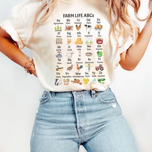 May include: A white t-shirt featuring a graphic of farm-related items and the text "FARM LIFE ABCS". Each item is paired with a letter of the alphabet and a word describing the item. For example, "Aa Agriculture" is paired with a small image of a plant.