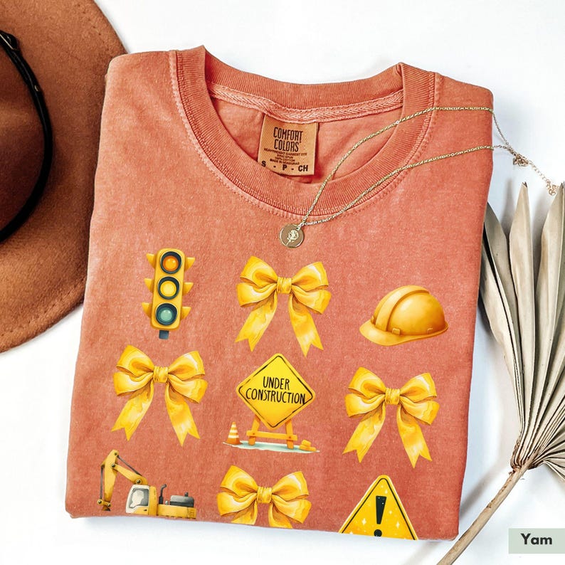 May include: A coral-colored t-shirt with a variety of yellow construction-themed illustrations, including a traffic light, a hard hat, a construction sign that says "Under Construction", and bows.