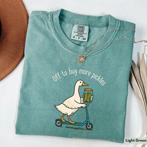 May include: Light green t-shirt with the slogan "Off to buy more pickles" and a cartoon duck on a scooter carrying a jar of pickles. The shirt has a crew neck and short sleeves, made by Comfort Colors.
