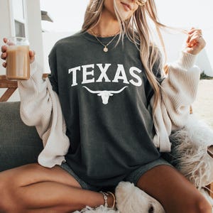 Puede incluir: Una camiseta gris oscuro con la palabra "TEXAS" impresa en blanco con una silueta blanca de un toro longhorn debajo.