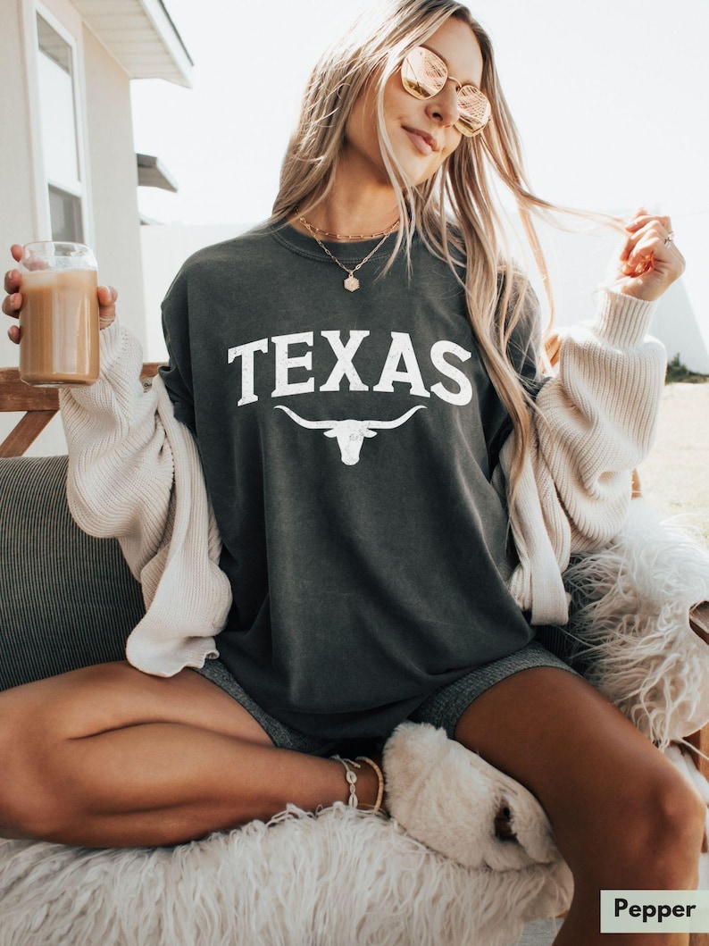 Puede incluir: Una camiseta gris oscuro con la palabra "TEXAS" impresa en blanco con una silueta blanca de un toro longhorn debajo.