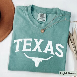 Puede incluir: Camiseta verde claro con la palabra "TEXAS" en letras blancas y una silueta blanca de un toro longhorn. La camiseta tiene un aspecto desgastado y est&aacute; hecha por Comfort Colors.