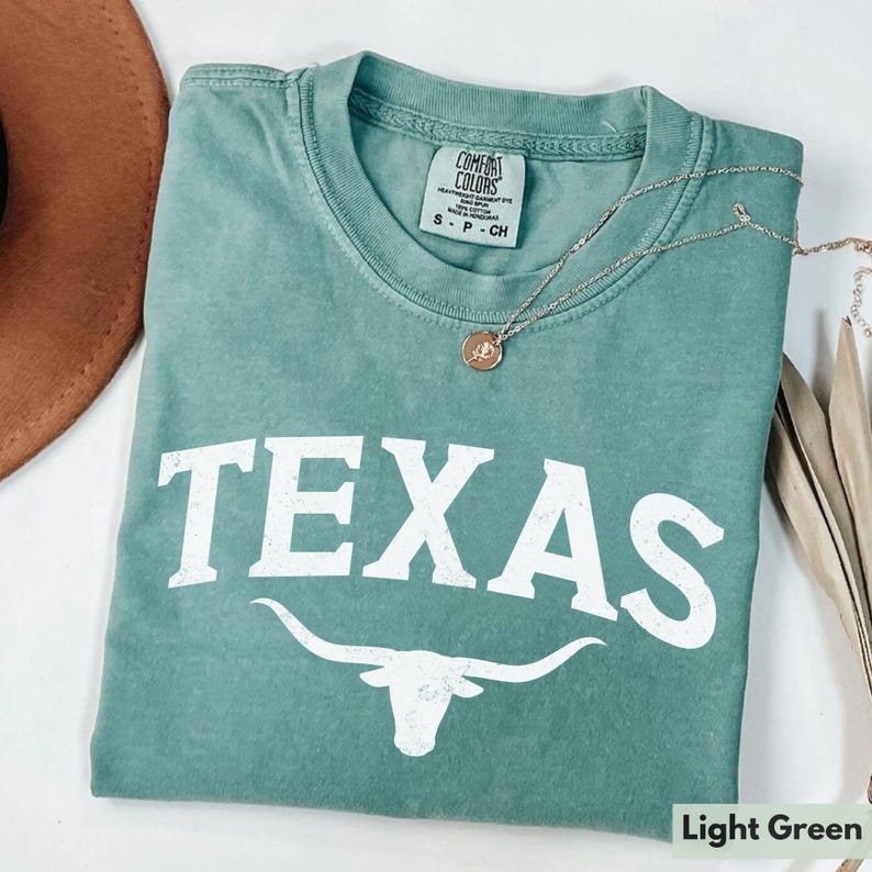 Puede incluir: Camiseta verde claro con la palabra "TEXAS" en letras blancas y una silueta blanca de un toro longhorn. La camiseta tiene un aspecto desgastado y est&aacute; hecha por Comfort Colors.