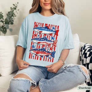 May include: Light blue t-shirt featuring a patriotic design with three fish in red, white, and blue. The fish are set against a background of red stripes and blue stars, evoking an American flag theme. The shirt is made of a soft material.