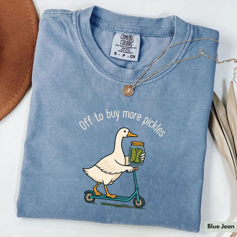 May include: A blue jean coloured t-shirt featuring a cartoon duck on a scooter, carrying a jar of pickles. The text "Off to buy more pickles" is above the duck. A gold necklace is draped across the shirt.