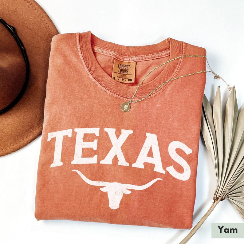 Puede incluir: Una camiseta naranja quemada con la palabra "TEXAS" impresa en blanco con una silueta blanca de un toro longhorn debajo. La camiseta est&aacute; doblada y colocada sobre una superficie blanca.