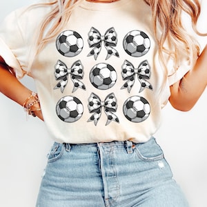 Puede incluir: Una camiseta blanca con un dise&ntilde;o gr&aacute;fico de balones de f&uacute;tbol blanco y negro y lazos. La camiseta est&aacute; en una modelo que lleva pantalones vaqueros azules.
