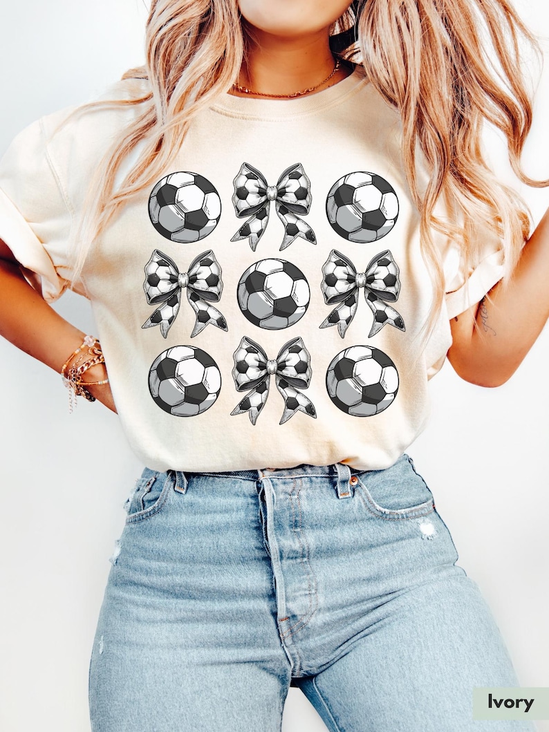 Puede incluir: Una camiseta blanca con un dise&ntilde;o gr&aacute;fico de balones de f&uacute;tbol blanco y negro y lazos. La camiseta est&aacute; en una modelo que lleva pantalones vaqueros azules.