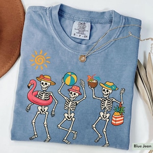 May include: A blue jean t-shirt with a graphic of three skeletons on a beach. The skeletons are wearing hats and sunglasses and are holding beach items like a beach ball, a pineapple, and a coconut. The text "Comfort Colors" is visible on the tag.