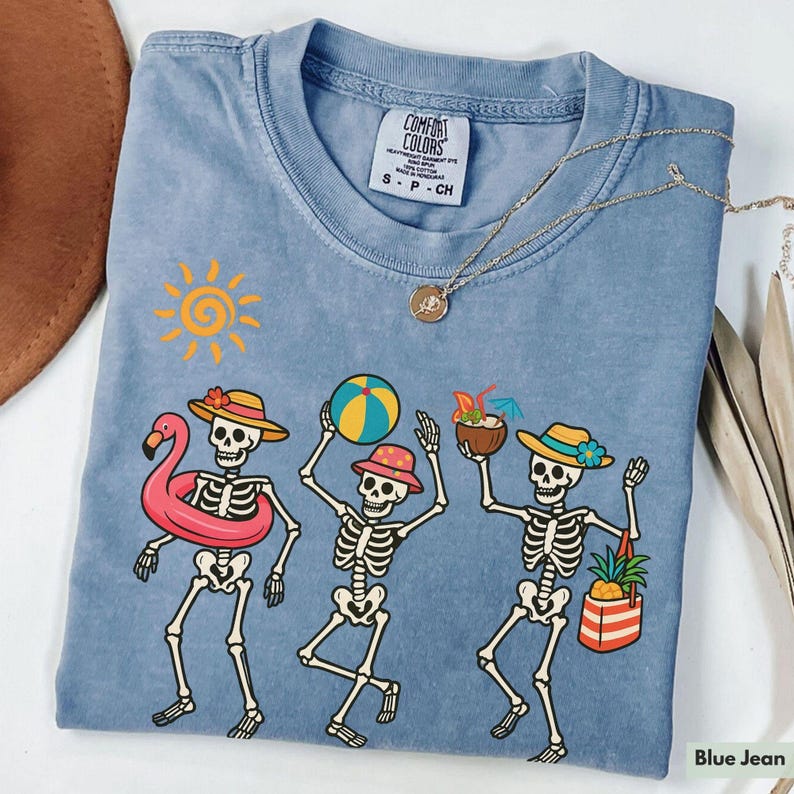 May include: A blue jean t-shirt with a graphic of three skeletons on a beach. The skeletons are wearing hats and sunglasses and are holding beach items like a beach ball, a pineapple, and a coconut. The text "Comfort Colors" is visible on the tag.