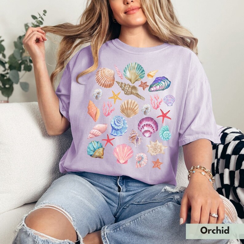 Op de afbeelding: Een lavendelkleurig T-shirt met een aquarelontwerp van verschillende schelpen, zeesterren en koralen. Het ontwerp is gecentreerd op het shirt en heeft een verscheidenheid aan kleuren, waaronder roze, blauw, groen en geel.