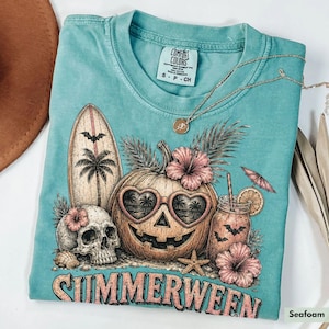May include: Seafoam green t-shirt with a graphic design featuring a pumpkin with heart-shaped sunglasses, a skull, a surfboard, and a cocktail. The word "SUMMERWEEN" is printed at the bottom. A Comfort Colors tag is visible at the top.