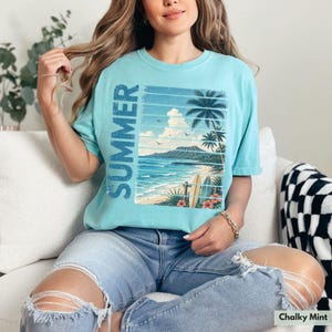 May include: A light blue t-shirt with a beach scene graphic and the word "SUMMER" printed vertically. The graphic features a beach, ocean, palm trees, and surfboards. The shirt is a relaxed fit and is made of a soft material.