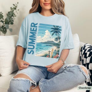 May include: Light blue t-shirt with a beach scene graphic and the word "SUMMER" in vertical blue text. The graphic features a beach with palm trees, surfboards, and ocean waves. The shirt is made of a soft material.