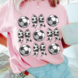 Puede incluir: Camiseta rosa con un dise&ntilde;o gr&aacute;fico de balones de f&uacute;tbol y lazos. El dise&ntilde;o se repite nueve veces en la camiseta.