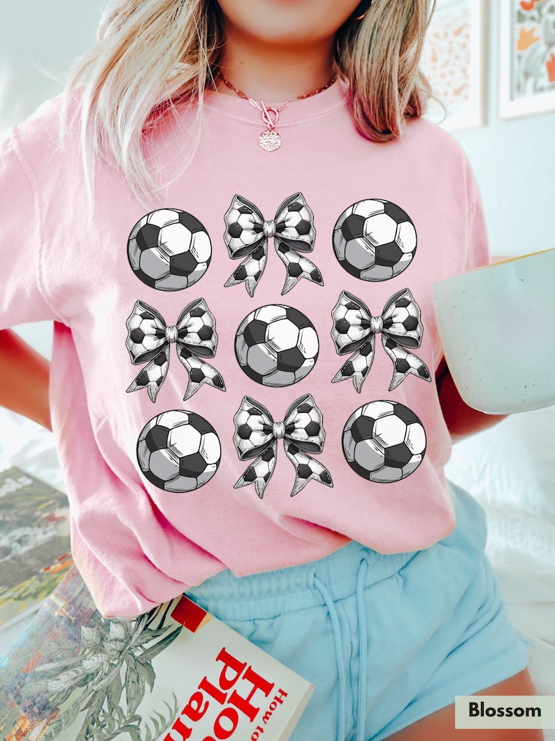 Puede incluir: Camiseta rosa con un dise&ntilde;o gr&aacute;fico de balones de f&uacute;tbol y lazos. El dise&ntilde;o se repite nueve veces en la camiseta.