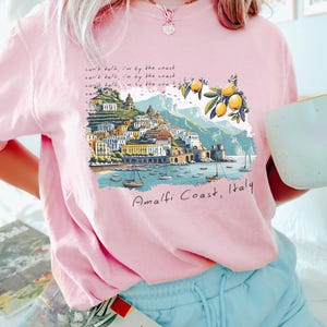以下が含まれることがあります： 海岸の風景が描かれたピンクのTシャツ。イタリアの amalfi 海岸の風景で、レモンの木とボートが描かれています。Tシャツには「can't talk, I'm by the coast.」という文字が書かれています。