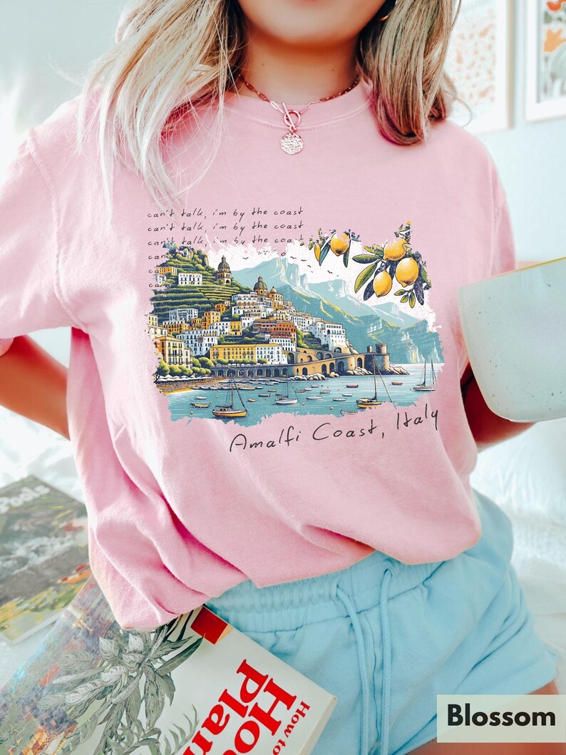 以下が含まれることがあります： 海岸の風景が描かれたピンクのTシャツ。イタリアの amalfi 海岸の風景で、レモンの木とボートが描かれています。Tシャツには「can't talk, I'm by the coast.」という文字が書かれています。