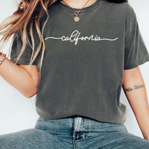 May include: A dark gray t-shirt with the word "california" written in white cursive script. The model is wearing the shirt with blue jeans and a gold necklace.