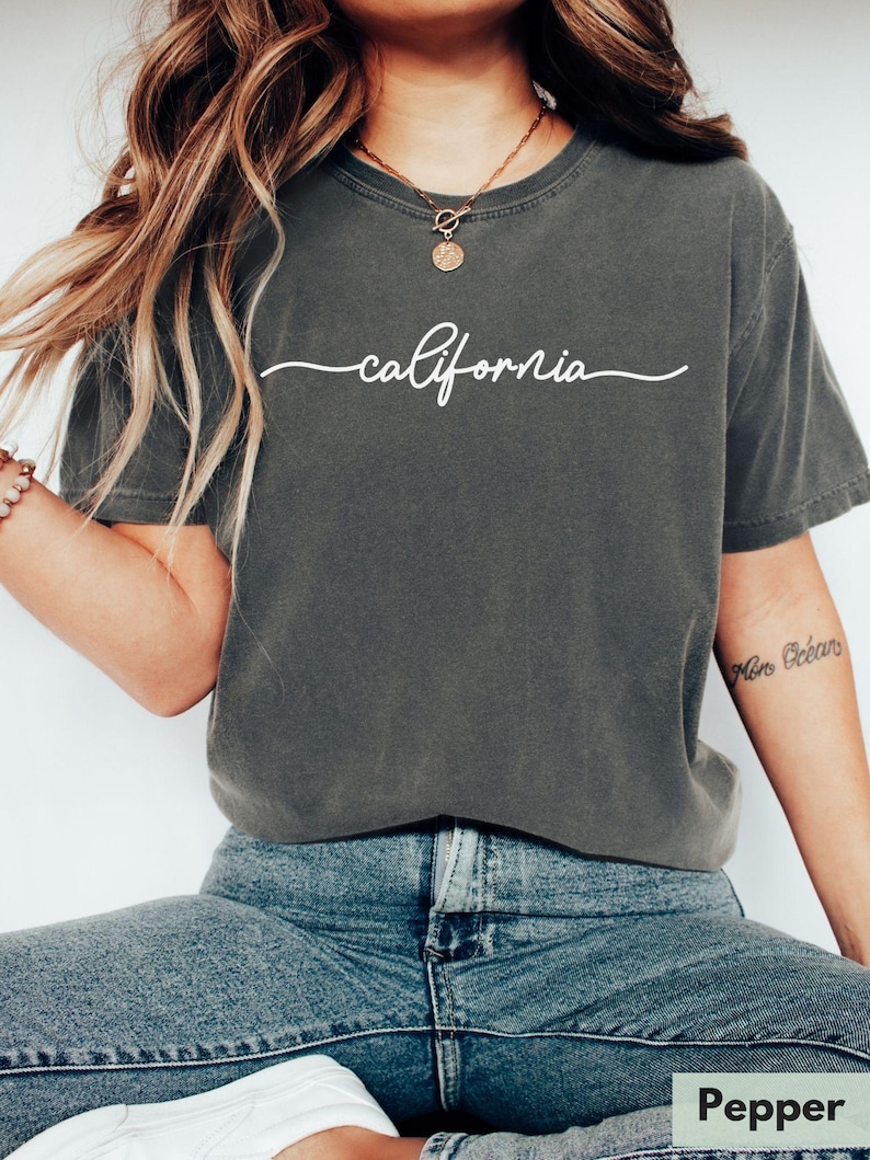 May include: A dark gray t-shirt with the word "california" written in white cursive script. The model is wearing the shirt with blue jeans and a gold necklace.