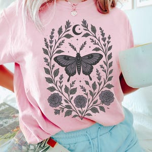 May include: A pink t-shirt with a black butterfly graphic surrounded by floral designs and a crescent moon. The butterfly has a cross on its body. The shirt has a crew neck and short sleeves.
