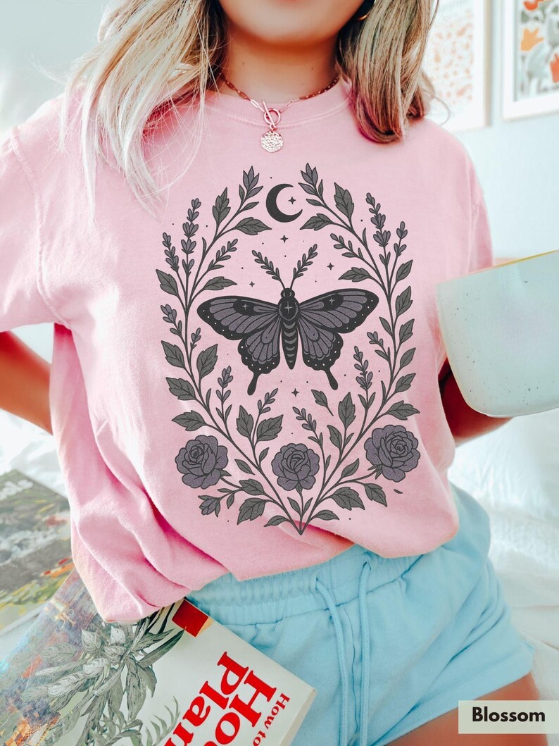 May include: A pink t-shirt with a black butterfly graphic surrounded by floral designs and a crescent moon. The butterfly has a cross on its body. The shirt has a crew neck and short sleeves.