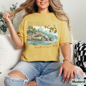 以下が含まれることがあります： マスタードイエローのTシャツに、イタリアのアマルフィ海岸のグラフィックがプリントされています。テキストには「can't talk, I'm by the coast」と「Amalfi Coast, Italy」が書かれています。