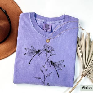 May include: A lavender colored t-shirt with a black dragonfly and flower design. The shirt has a tag that says "Comfort Colors".