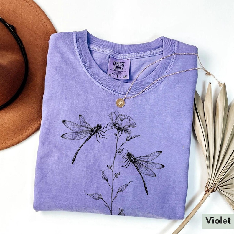 May include: A lavender colored t-shirt with a black dragonfly and flower design. The shirt has a tag that says "Comfort Colors".