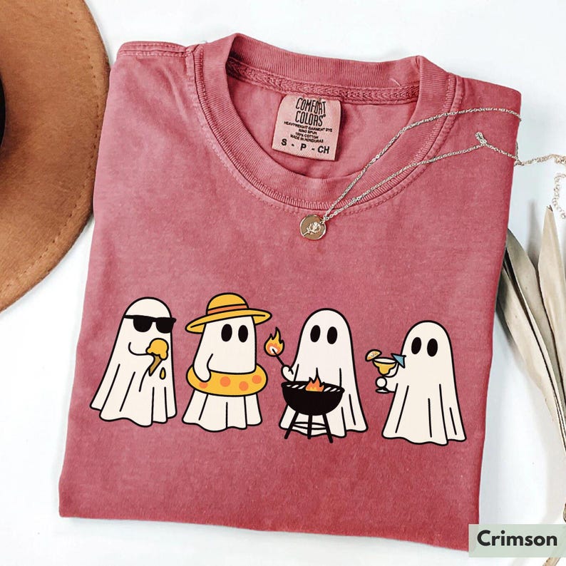 May include: Crimson-colored t-shirt featuring a graphic of four cartoon ghosts enjoying summer activities. One ghost wears sunglasses and eats ice cream, another wears a hat and floatie, one grills, and the last one sips a cocktail. The shirt has a Comfort Colors tag.