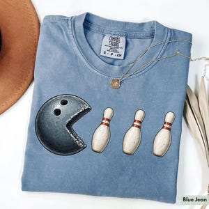 Op de afbeelding: Blauw spijkerbroekkleurig T-shirt met een afbeelding van een bowlingbal die kegels 'eet'. Het shirt heeft een ronde hals en korte mouwen. Het merklabel zegt "Comfort Colors". Een gouden ketting ligt over het shirt.