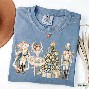 May include: A blue jean colored t-shirt featuring a graphic print of a mouse king, a ballerina, a decorated Christmas tree, and a nutcracker. The shirt has a Comfort Colors label at the neck and is displayed with a gold necklace and a brown hat.
