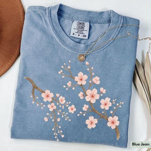 May include: A blue jean t-shirt with a floral design of pink cherry blossoms on a branch. The shirt has a tag that reads "Comfort Colors" and "S - P - CH".