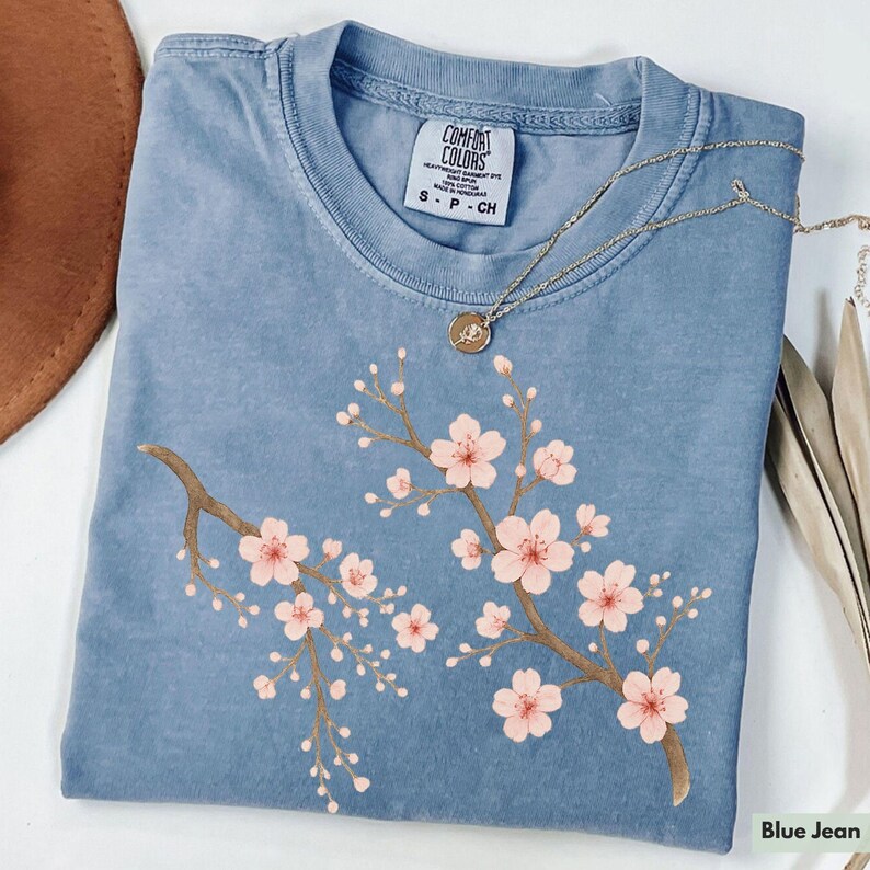 May include: A blue jean t-shirt with a floral design of pink cherry blossoms on a branch. The shirt has a tag that reads "Comfort Colors" and "S - P - CH".