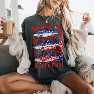 May include: Dark gray t-shirt featuring three fish designs with red, white, and blue colors, stars, and stripes. The fish are arranged vertically on the shirt. The word "Pepper" is in the bottom right corner.