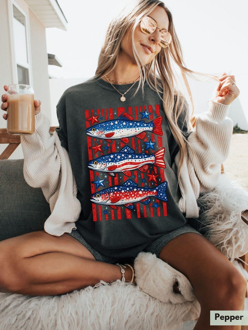 May include: Dark gray t-shirt featuring three fish designs with red, white, and blue colors, stars, and stripes. The fish are arranged vertically on the shirt. The word "Pepper" is in the bottom right corner.